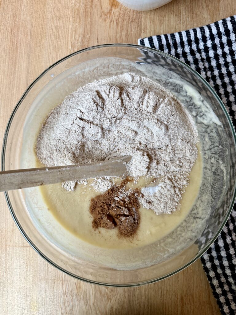 healthy banana bread recipe. Image of whole wheat flour, cinnamon, salt added to wet ingredients in glass bowl next to black and white dish cloth.