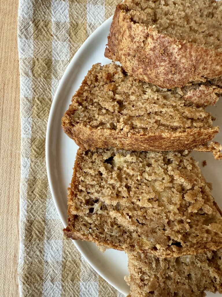 sourdough banana bread recipe. Image of 4 slices of banana bread, stacked on a white ceramic plate, sitting on a brown and white gingham cloth napkin on a wooden counter.