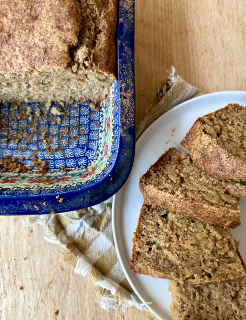 Image of banana bread in blue pattern loaf pan next to white plate with sliced banana bread sliced out on it. Pan and plate are on wooden counter.